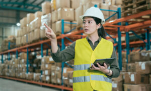 warehouse worker woman in safety vests holding digital tablet and pointing up with finger. young girl staff wearing hard hat working in stockroom. elegant lady employee taking inventory in storehouse